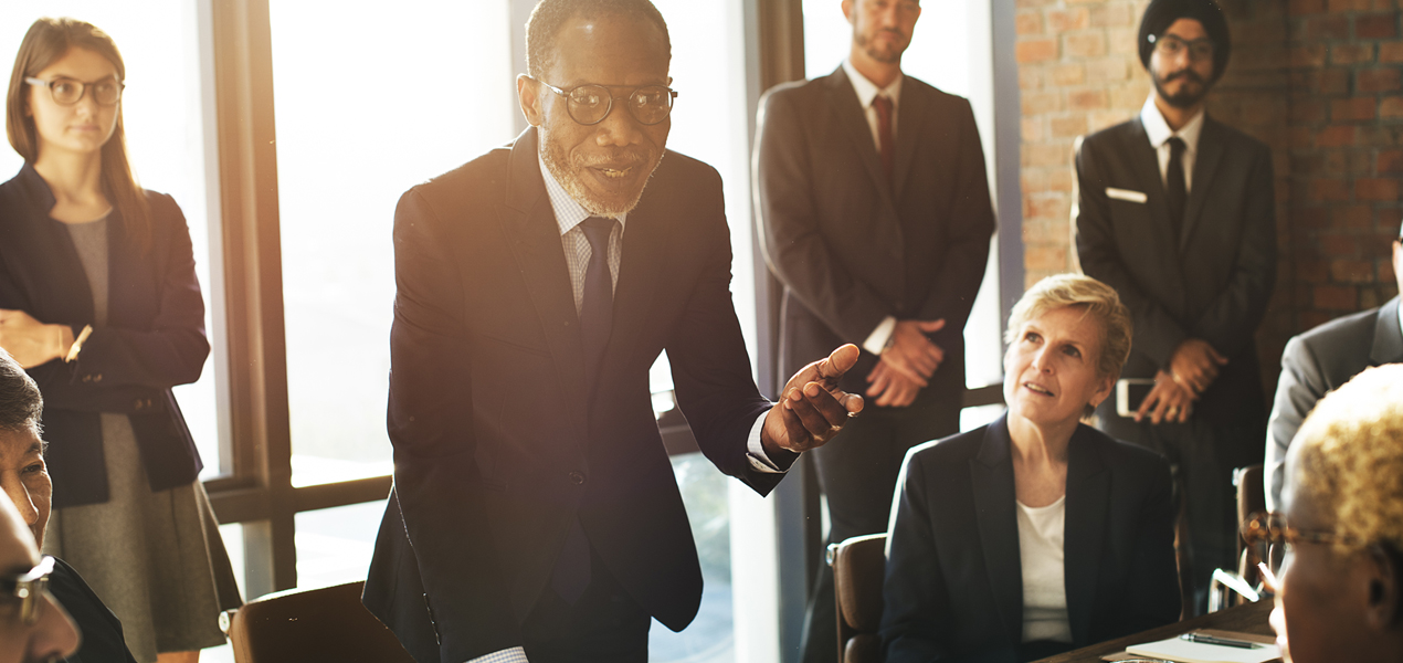 A team looking towards a man in a suit standing up and speaking during a meeting.