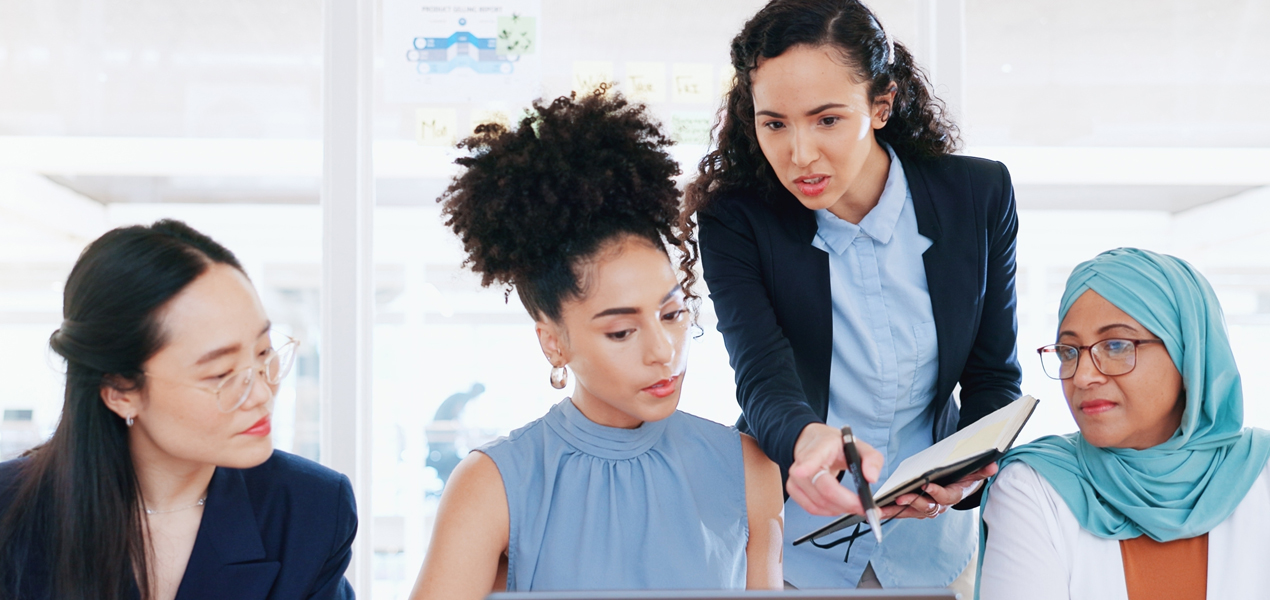 A group of women in business attire collaboratively reviewing information on a laptop.